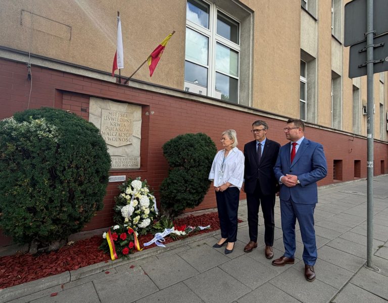 Na zewnątrz budynku. Dwóch mężczyzn i kobieta stoją pod tablicą pamiątkową, pod którą złożono wieńce.