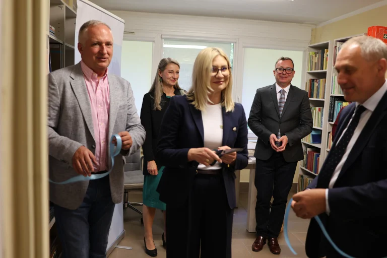 Grupa ludzi stoi przy wstędze. Kobieta trzyma w dłoni nożyce.