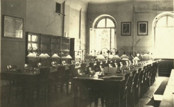 Czarno-biała fotografia przestronnej, pustej czytelni wyposażonej w długie stoły, drewniane krzesła oraz rzędy klasycznych lamp biurkowych.