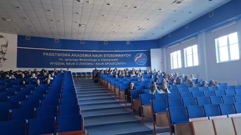 Aula wykładowa. Krzesła obite niebieską tkaniną, studenci siedzą zgromadzeni w dalszych rzędach.