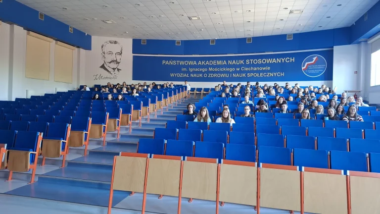 Aula wykładowa. Krzesła obite niebieską tkaniną, studenci siedzą zgromadzeni w dalszych rzędach.