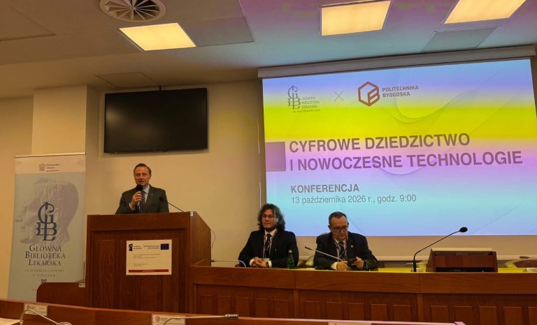 Mężczyzna w garniturze przemawia do mikrofonu, stojąc za drewnianą mównicą. Obok niego, przy stole konferencyjnym, siedzą dwaj mężczyźni. W tle widać slajd tytułowy konferencji „Cyfrowe Dziedzictwo i Nowoczesne Technologie”.