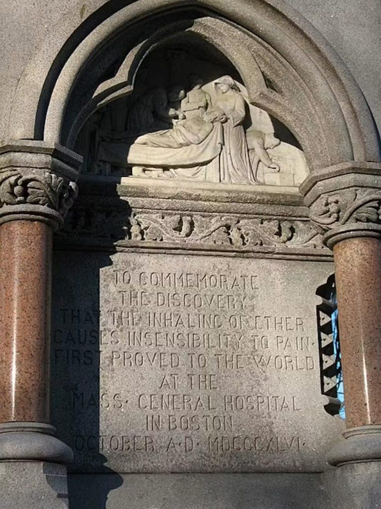 Fragment kamiennego pomnika w Bostonie. W łuku znajduje się płaskorzeźba przedstawiająca postacie pochylone nad pacjentem, a poniżej wyryto angielską inskrypcję upamiętniającą odkrycie znieczulenia eterem w 1846 roku.