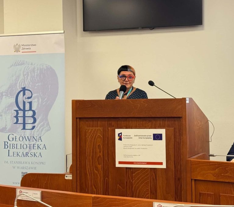 Kobieta w okularach i ciemnej bluzce w kropki przemawia przy drewnianej mównicy w sali konferencyjnej. Obok mównicy stoi rolowana tablica informacyjna Głównej Biblioteki Lekarskiej im. Stanisława Konopki w Warszawie.