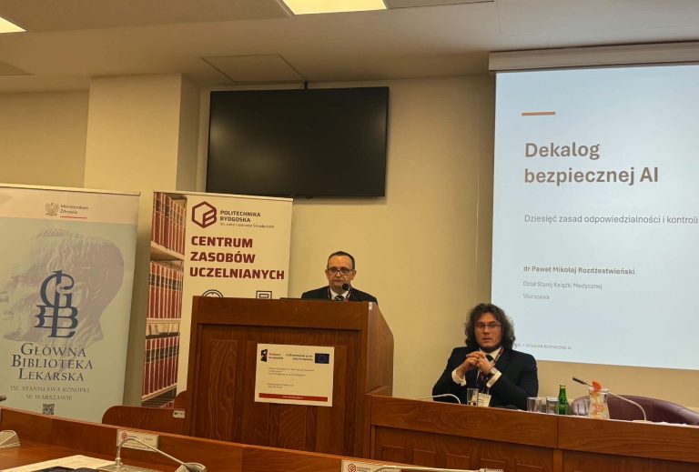 Mężczyzna w okularach i garniturze przemawia z za mównicy. Z boku wyświetlany jest slajd prezentacji pt. „Dekalog bezpiecznej AI. Dziesięć zasad odpowiedzialności i kontroli” autorstwa dr. Pawła Mikołaja Rozdżestwieńskiego. Przed nim przy stole siedzi inny uczestnik spotkania.