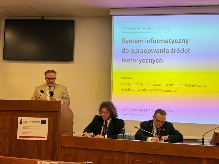 Mężczyzna w jasnej marynarce przemawia przy mównicy. Na ekranie obok widnieje slajd: „System informatyczny do opracowania źródeł historycznych. Od digitalizacji i rozpoznawania tekstu do semantycznej eksploracji wiedzy z wykorzystaniem AI” (autor: mgr inż. Jacek Suty). Przy stole obok dwóch mężczyzn pisze w notatnikach.