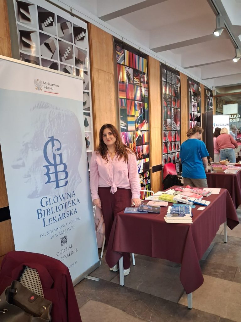 Kobieta w różowej bluzce stoi przy stanowisku - stole z książkami i ulotkami. Obok rollup z logo Głównej Biblioteki Lekarskiej.