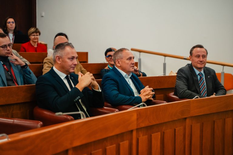 Trzech mężczyzn w garniturach siedzących za drewnianą ławką podczas wydarzenia w sali wykładowej.