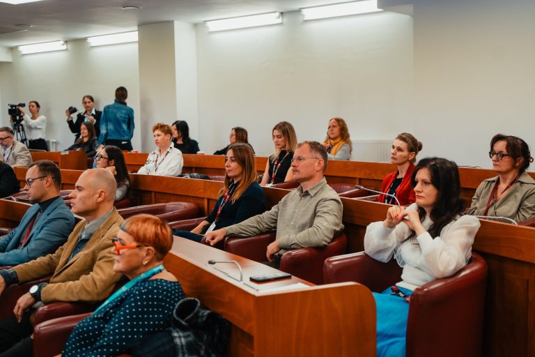 Uczestnicy konferencji siedzący w rzędach drewnianych ławek, słuchający prelekcji.