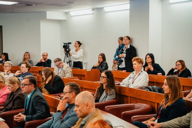 Uczestnicy konferencji siedzący w rzędach drewnianych ławek, słuchający prelekcji.