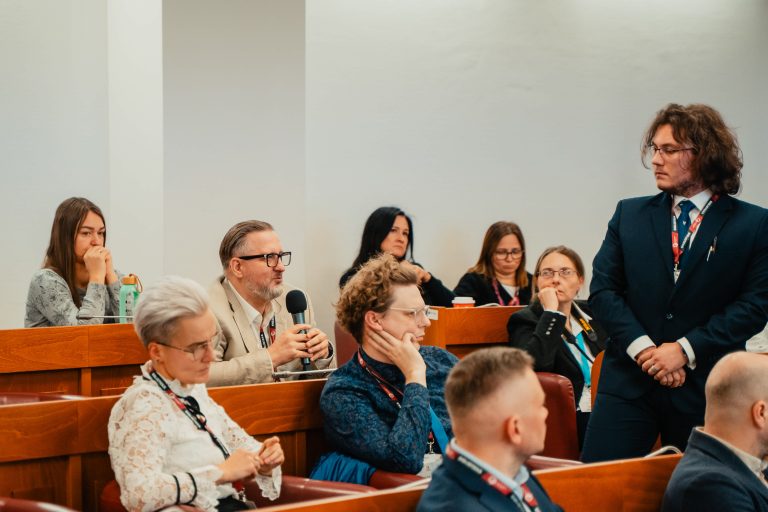 Uczestnicy konferencji siedzący w rzędach drewnianych ławek, słuchający prelekcji.