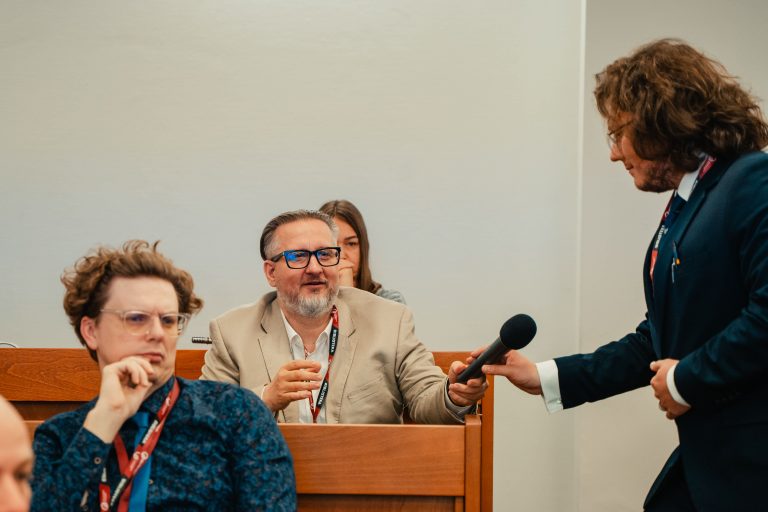 Mężczyzna stojący w przejściu podaje mikrofon uczestnikowi siedzącemu w ławce na widowni.