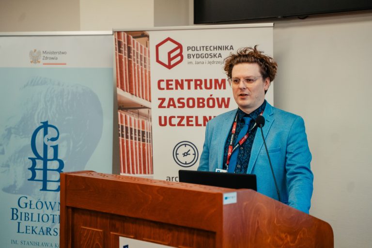 Prelegent w niebieskiej marynarce przemawiający zza drewnianej mównicy, w tle m.in. banner Politechniki Bydgoskiej.
