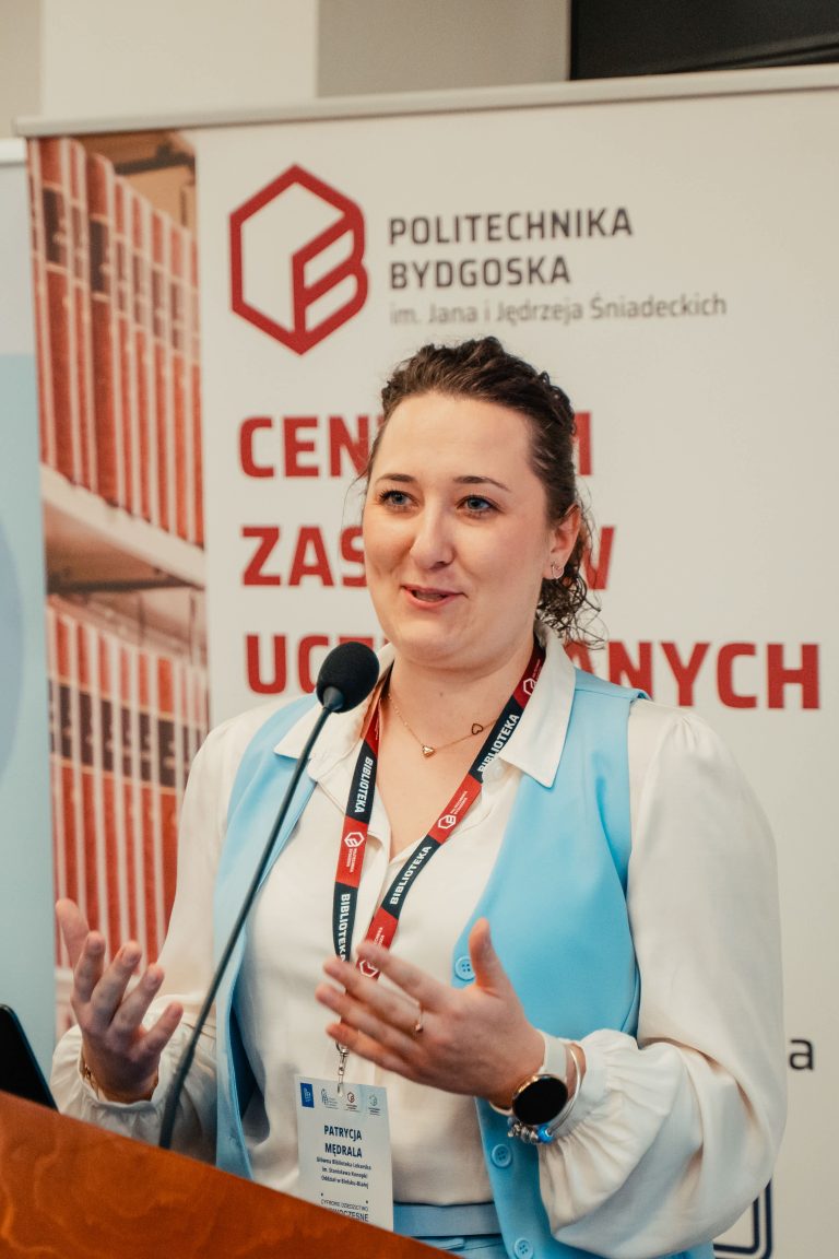 Kobieta w błękitnej kamizelce przemawiająca przez mikrofon zza mównicy na tle banneru Politechniki Bydgoskiej.