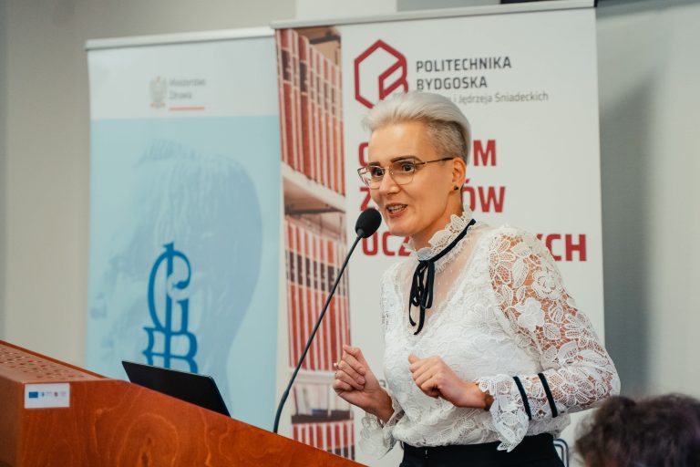 Kobieta w białej koronkowej bluzce przemawiająca przez mikrofon zza drewnianej mównicy na tle banneru Politechniki Bydgoskiej.